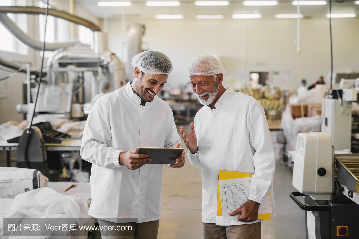 两位愉快的男同事穿着无菌的衣服,站在食品厂的电子平板电脑上看销售文件。